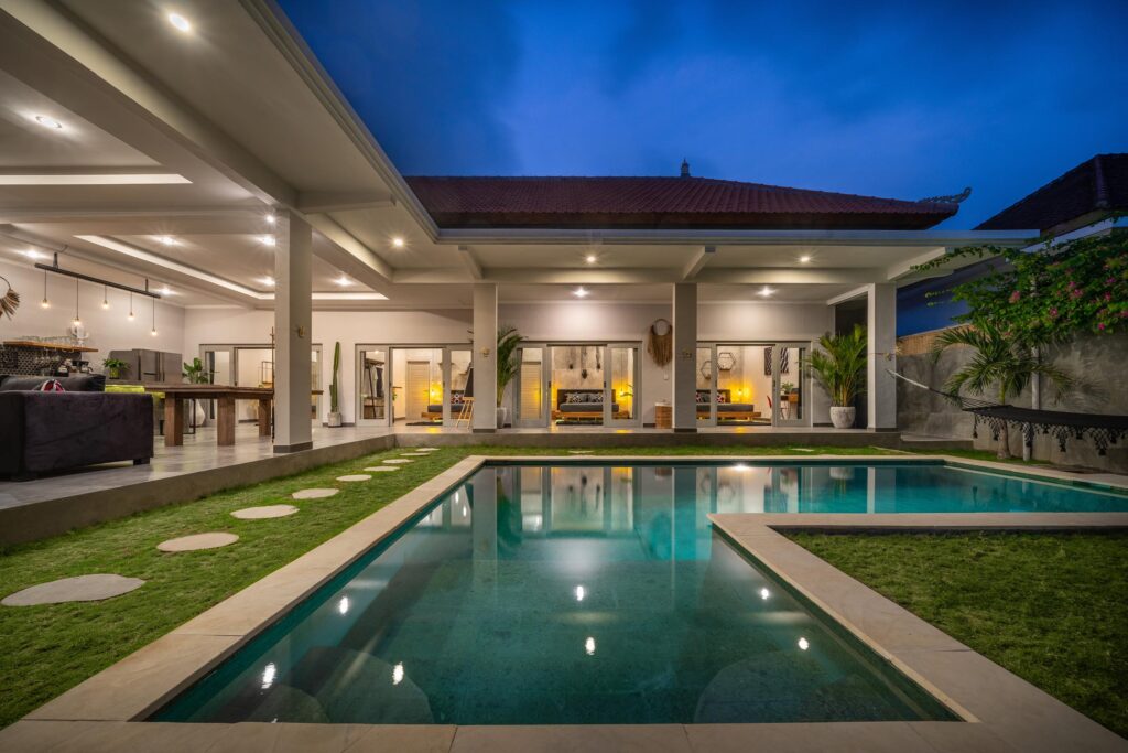 Modern villa with open-plan living area and illuminated swimming pool surrounded by grass and tropical plants at dusk.