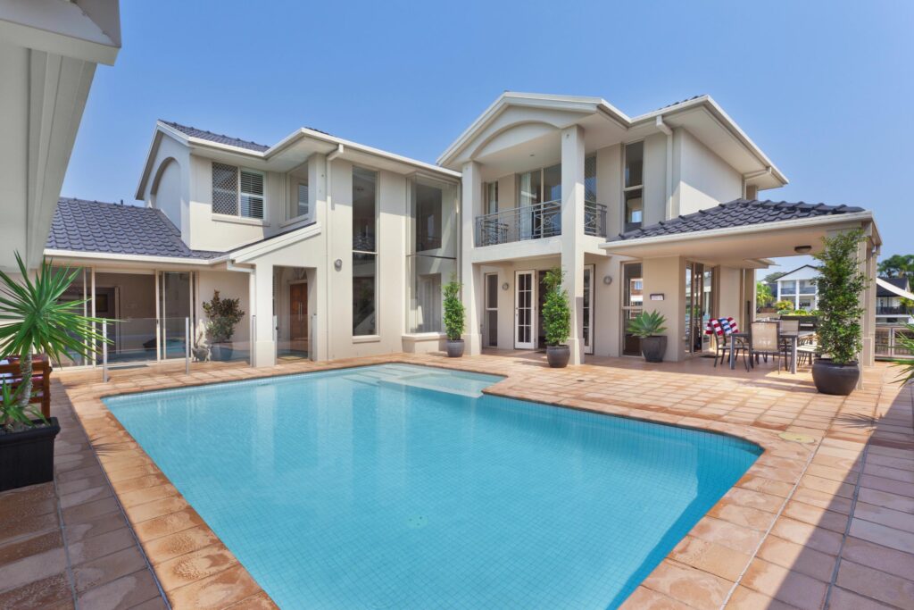 Modern two-story house with white exterior, tiled roof, and large windows, featuring a rectangular swimming pool surrounded by a tiled patio and potted plants. This is an example of the quality we bring to all our service areas.