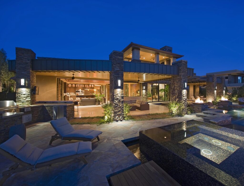 Modern luxury house at dusk with illuminated patio, outdoor seating, lounge chairs, and a pool with a built-in hot tub in the foreground.