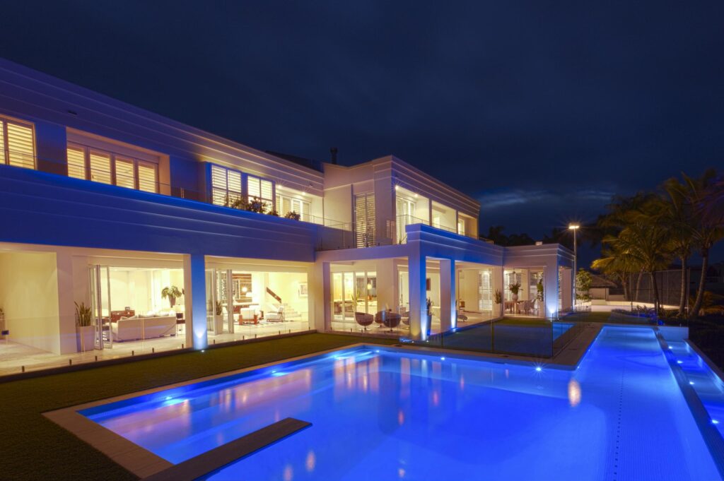 Clear Water Pools: Custom pool builders in Metro Atlanta: Modern two-story house with large windows and outdoor lighting, featuring a lit swimming pool at night. The interior is visible through the glass.
