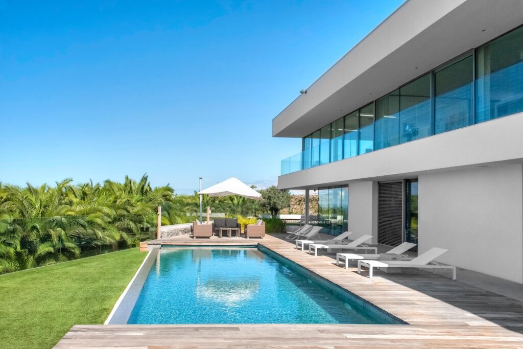 Modern house with large glass windows, patio, lounge chairs, swimming pool, and a seating area under a white umbrella, surrounded by green grass and palm trees.