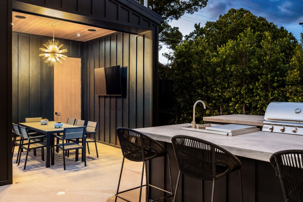 Modern outdoor patio featuring outdoor kitchens and fire features, a dining area with a table and chairs, a wall-mounted TV, and a geometric light fixture, all surrounded by lush greenery.