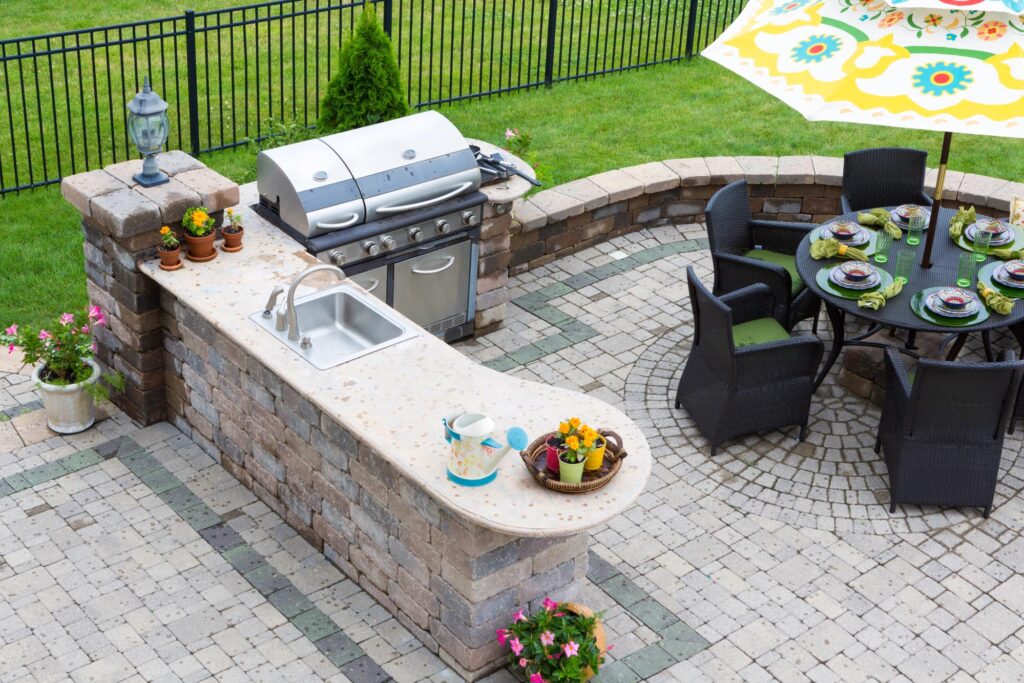 Outdoor patio with a built-in grill and sink, wicker dining set with plates, an umbrella, potted plants, and a stone paving pattern.