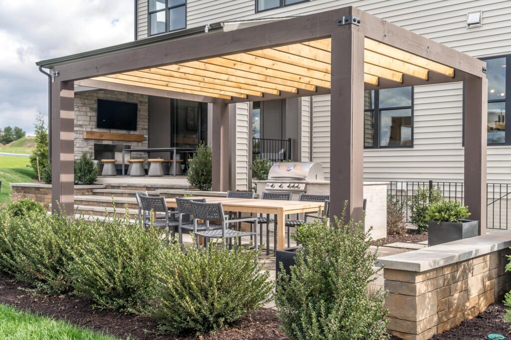 Modern outdoor patio with a wooden pergola, dining table, chairs, grill, television, and landscaped shrubs in front of a house.