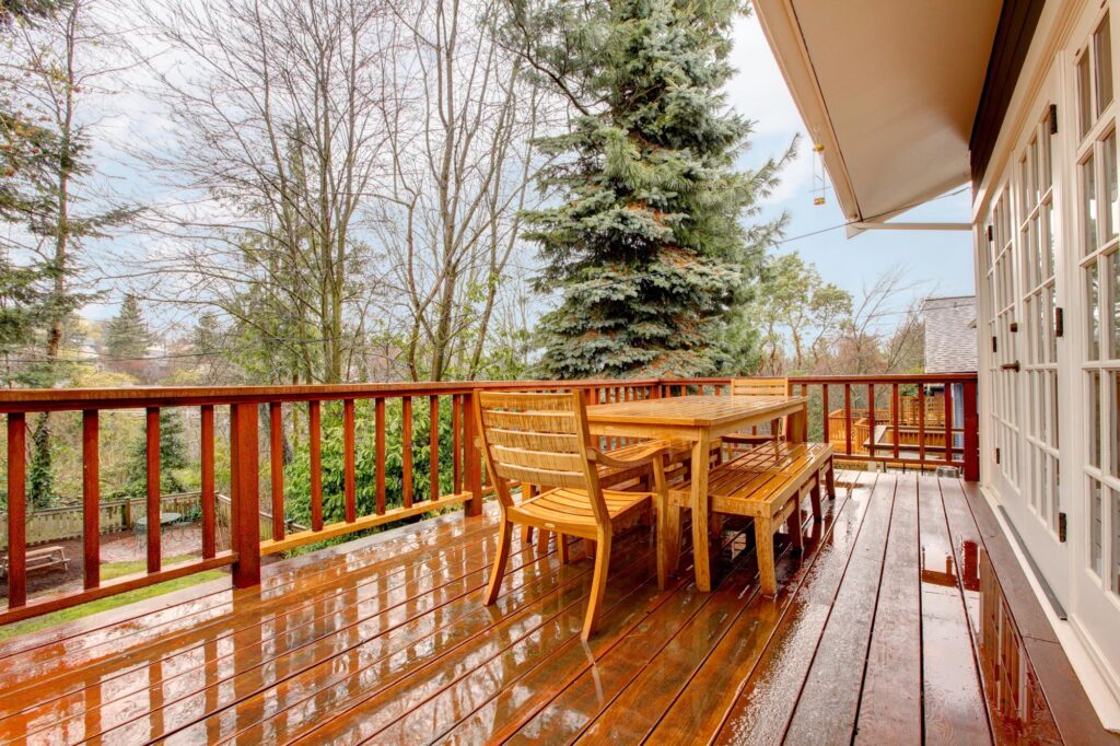 A wooden deck with a table and benches overlooks a yard with trees. The deck is wet, suggesting recent rain. Glass doors provide access to the indoor area.