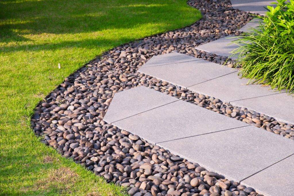 A curved garden pathway with large flat stone pavers, bordered by small river rocks, separates green grass from leafy plants.
