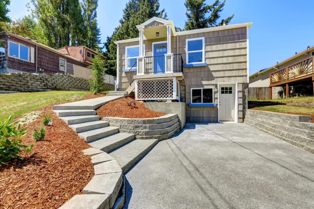 A small, beige house with a raised front porch, concrete driveway, curved steps, and landscaped yard with mulch and retaining walls.