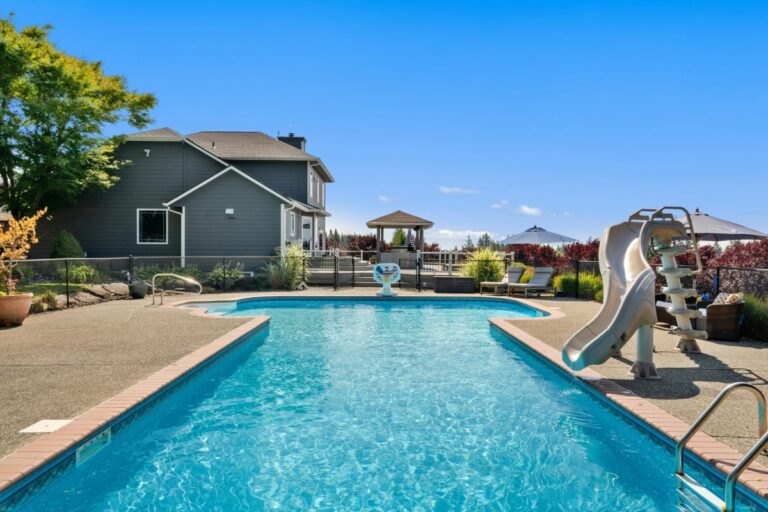 Rectangular outdoor swimming pool with a waterslide, surrounded by a concrete deck, next to a gray house and a gazebo under a clear blue sky—perfect for relaxing or practicing your favorite pool maintenance tips.
