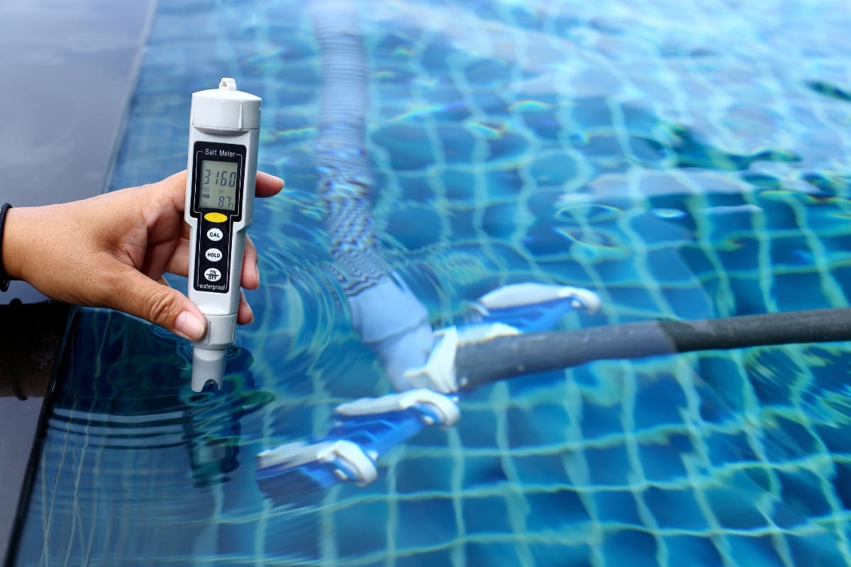 A hand holds a digital salt meter partially submerged in a swimming pool, displaying a reading, with a pool cleaning device visible underwater—a great example of practical pool maintenance tips in action.