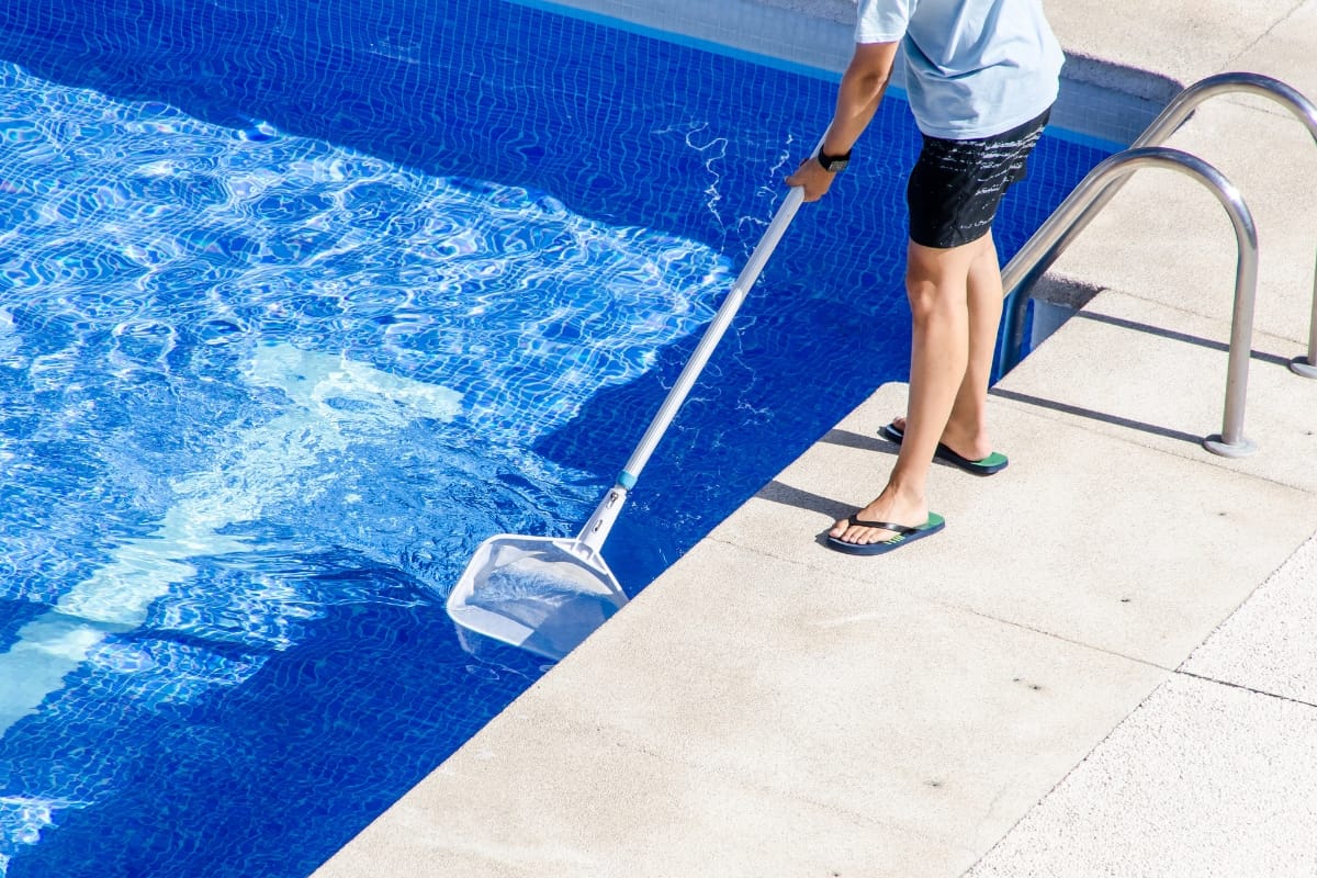 Person standing on poolside using a net skimmer to clean debris from the surface of a blue swimming pool, showcasing smart pool maintenance tips in action.