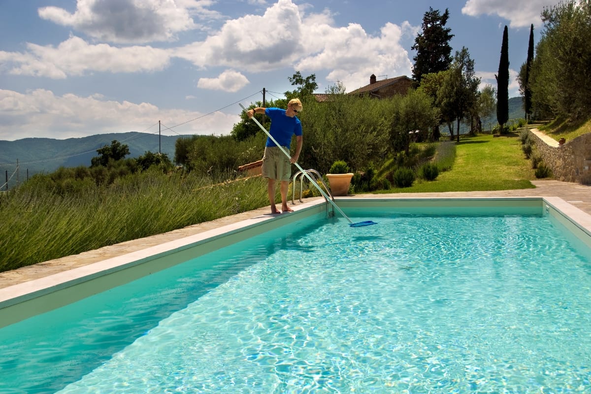 A person stands at the edge of an outdoor pool using a long pole to clean the water, demonstrating pool maintenance tips, with trees and hills visible in the background.