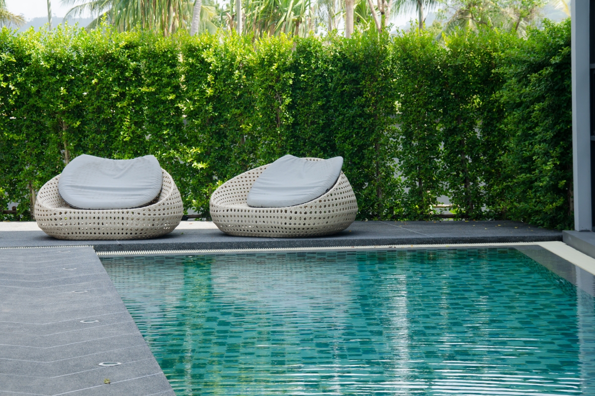 Two round wicker lounge chairs with white cushions sit beside a sparkling swimming pool, perfect for relaxing after a recent pool repair, with a tall green hedge and trees providing privacy in the background.