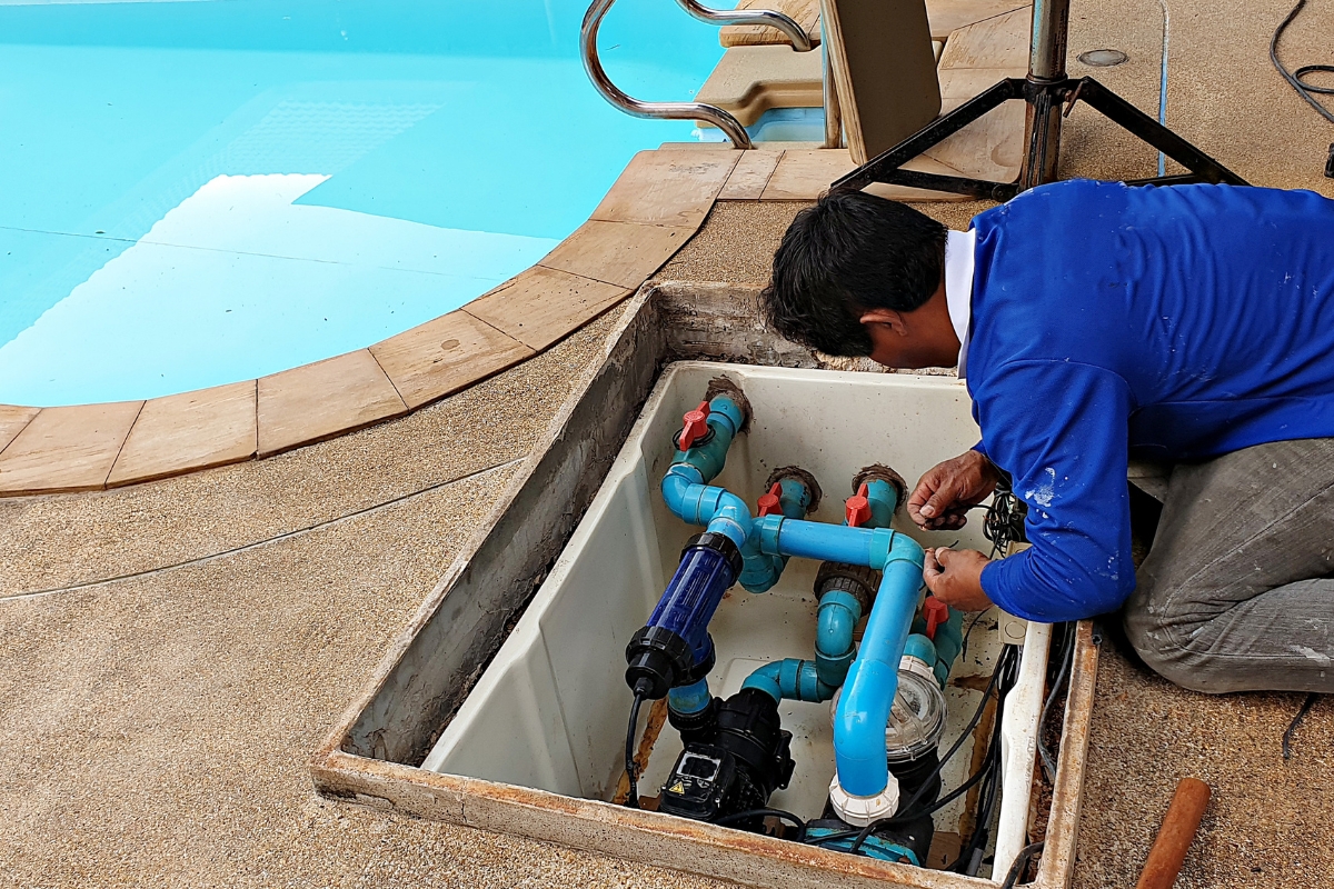 A worker performs pool repair on blue plumbing pipes in a maintenance hatch beside a swimming pool.