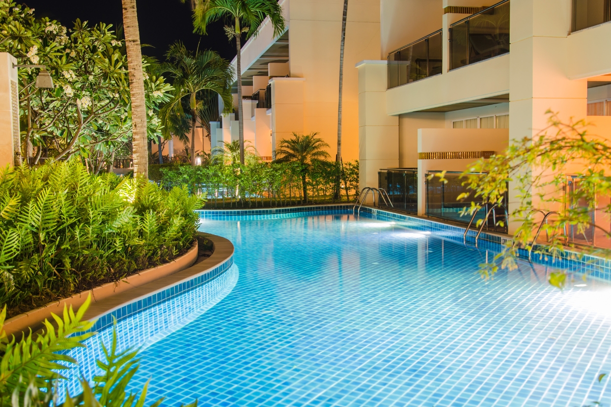 Outdoor swimming pool at night with calm blue water, surrounded by tropical plants and modern hotel buildings lit by warm lights—expert pool repair ensures the serene ambiance is always inviting.