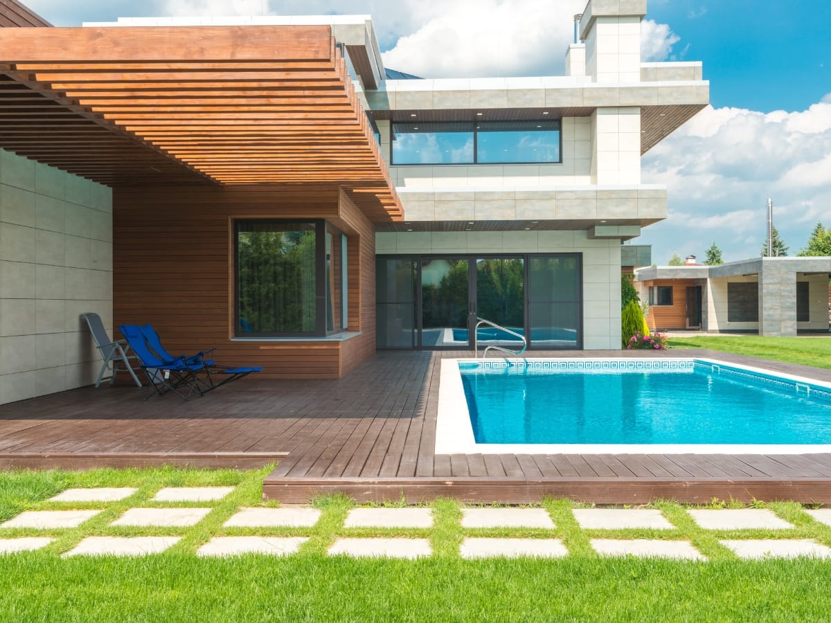 Modern house with large windows, wooden deck, two blue lounge chairs, and a stylish above-ground pool, surrounded by green lawn and stone pavers on a sunny day.