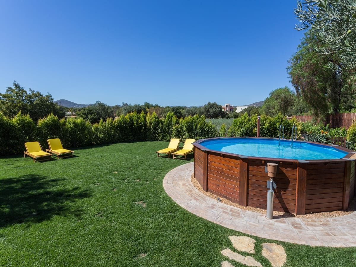 Above-ground pool with clear blue water sits in a grassy backyard, surrounded by trees and bushes, with four yellow lounge chairs inviting you to relax on the lawn.