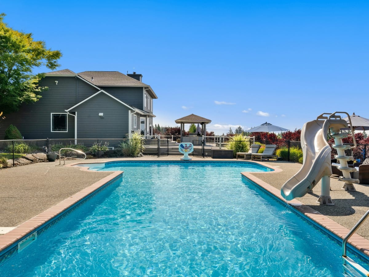 A backyard above-ground pool with a slide, surrounded by a patio area with lounge chairs, greenery, and a gazebo near a two-story gray house under a clear blue sky.