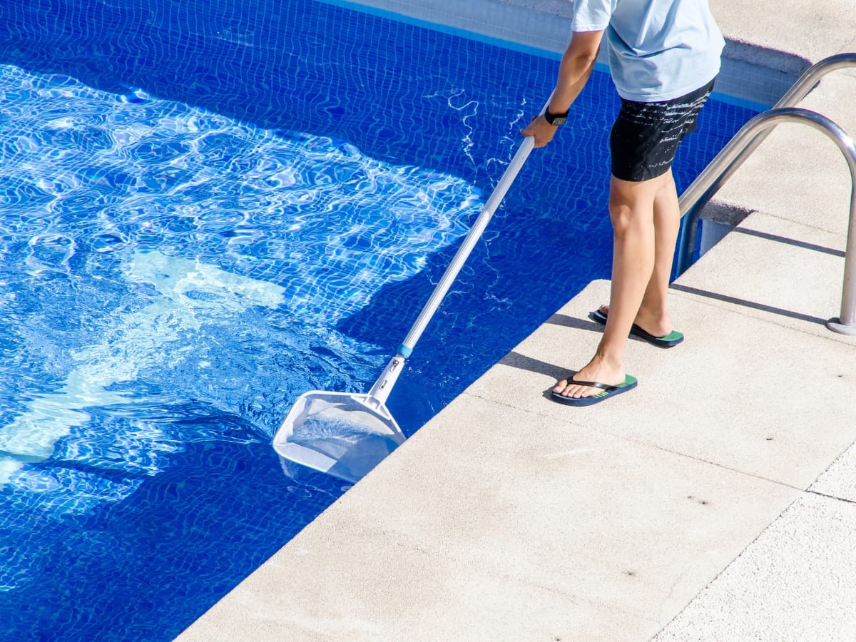Person standing at the edge of an above-ground pool uses a long pole with a net to clean the water.