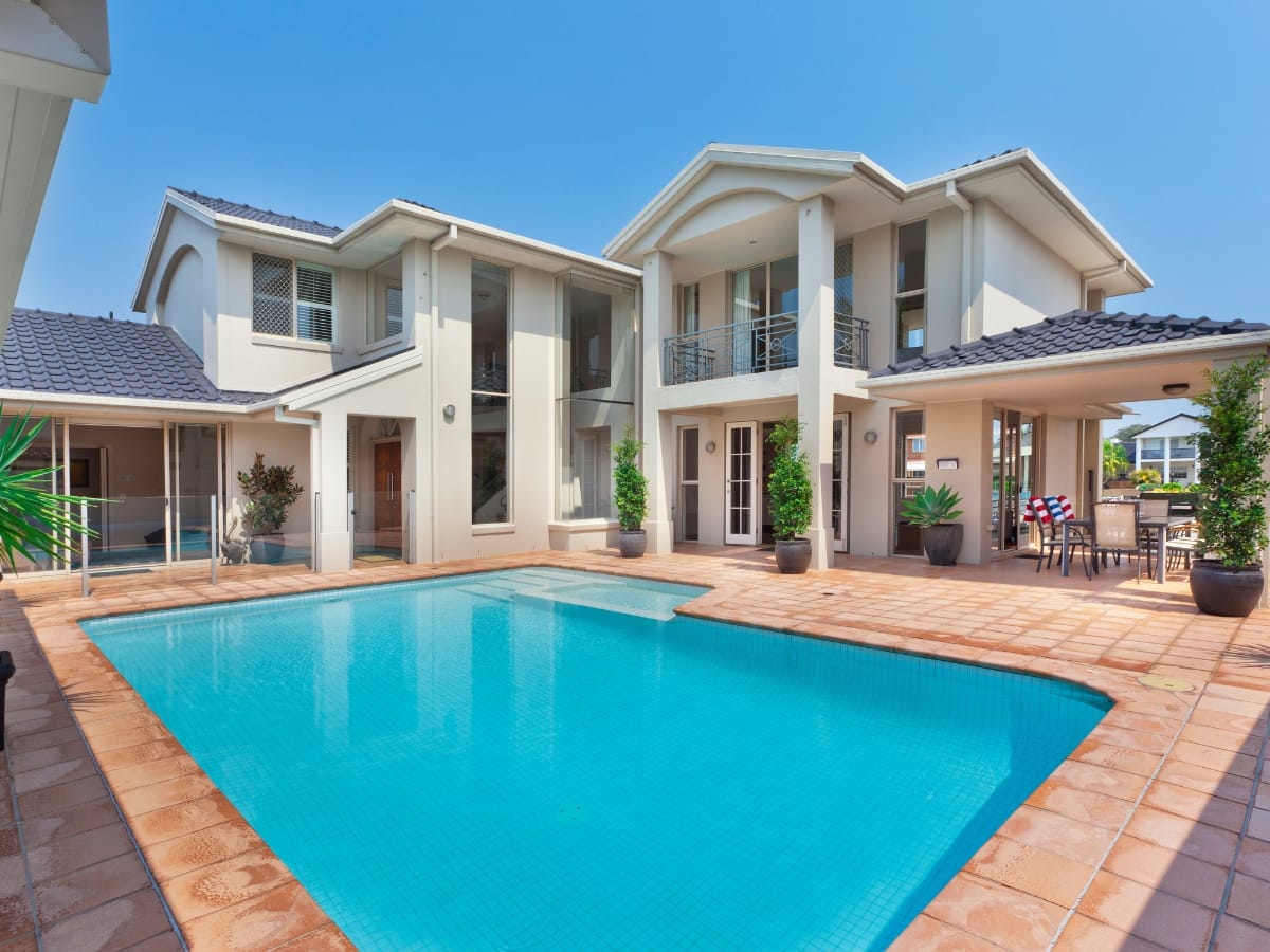 Modern two-story house with beige walls and blue roof tiles, featuring an above-ground pool and tiled patio under a clear sky.