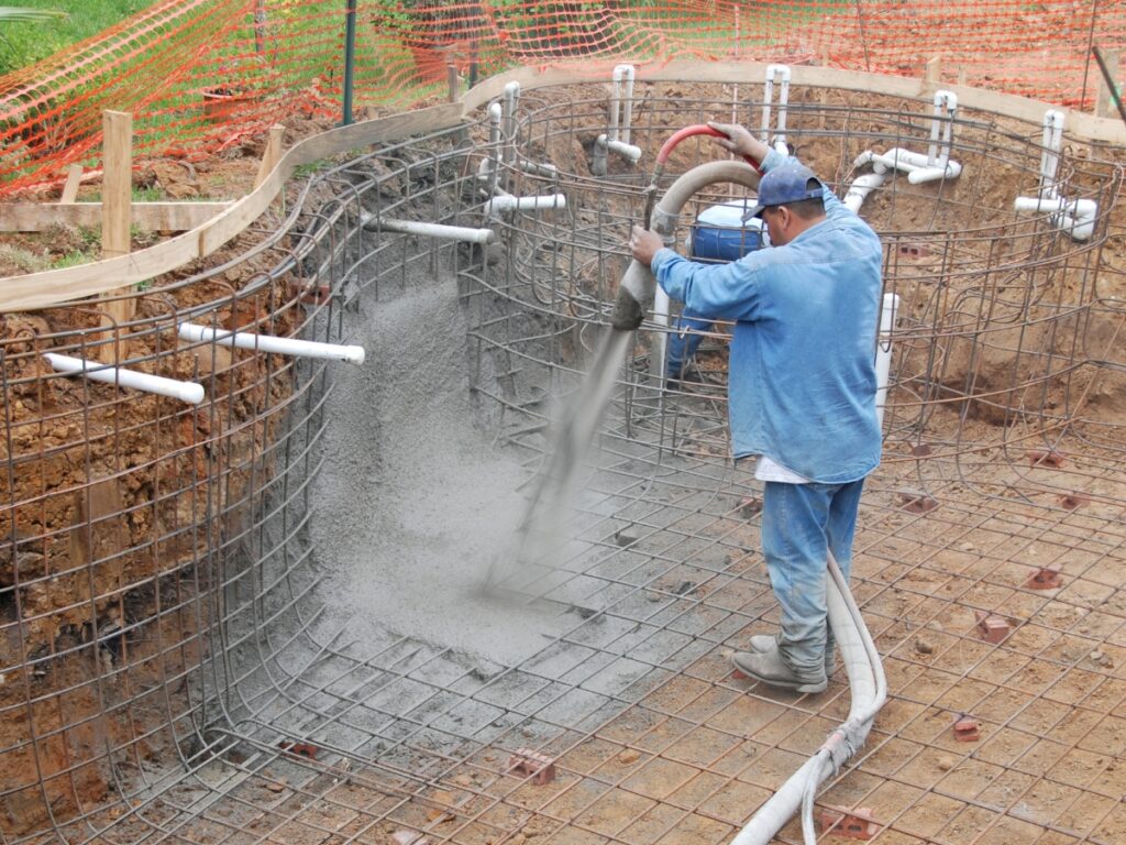 A worker sprays concrete onto a steel rebar frame for an in-ground pool renovation in Dawsonville, with pipes and orange safety fencing visible.