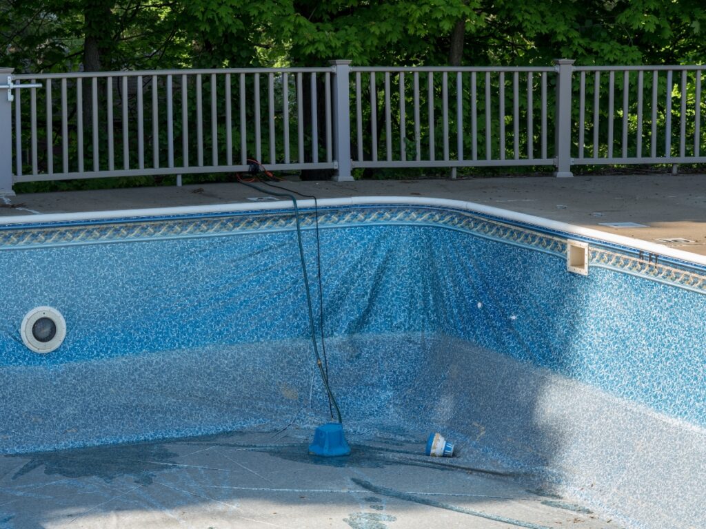 Empty, partially lined in-ground swimming pool with some debris and equipment at the bottom; a fence and trees are visible in the background—an ideal scene to consider pool renovation Dawsonville during the best time for upgrades.
