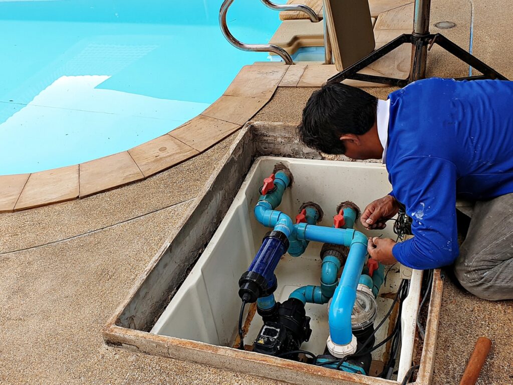 A person in a blue shirt repairs pool plumbing equipment inside an open maintenance hatch next to a swimming pool in Dawsonville, ensuring the best timing for pool renovation and maintenance.