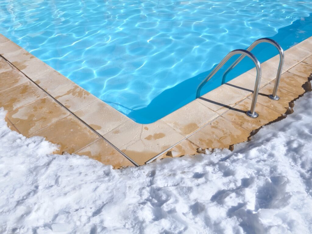 A swimming pool with metal steps sits beside a snow-covered deck, illustrating the need to winterize your pool as summer and winter elements collide.