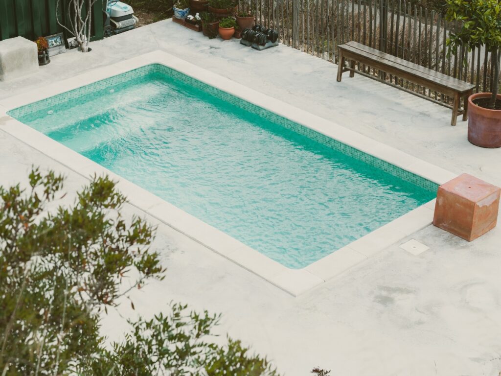 A rectangular swimming pool with clear water is surrounded by concrete, potted plants, a wooden bench, and a few outdoor objects—reminding you it’s important to winterize your pool for protection during colder months.
