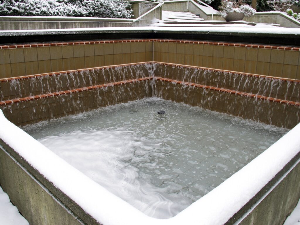 A square outdoor fountain with water cascading over tiled edges, surrounded by snow on the ledges and steps in the background—reminding you it’s time to winterize your pool for the season.