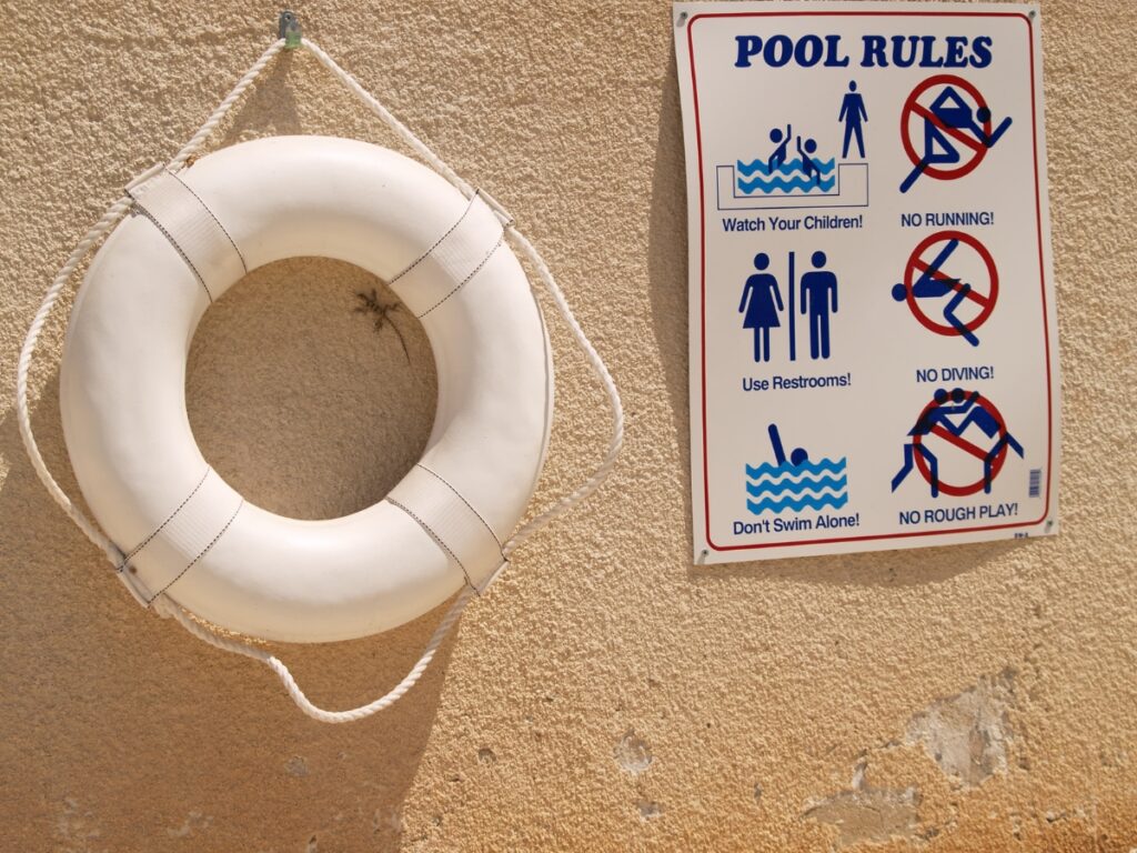 A life preserver hangs on a textured beige wall next to a sign listing pool rules, helping to prevent the Most Common Pool Safety Mistakes like running, diving, or rough play.