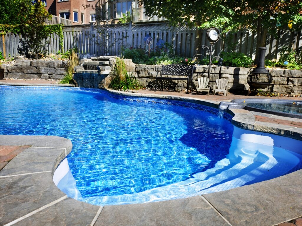 A backyard swimming pool with clear blue water, surrounded by stone edging, a small waterfall, garden landscaping, and patio seating—just be mindful of the most common pool safety mistakes to keep your oasis both beautiful and safe.