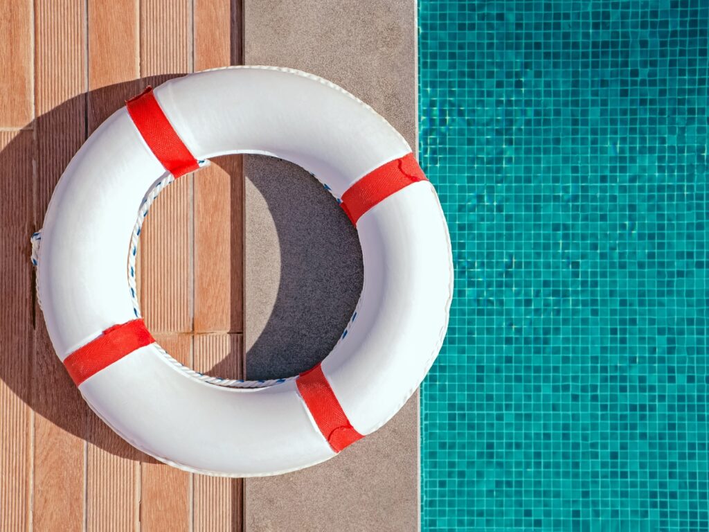 A white life preserver with red bands sits on the edge of a swimming pool with blue mosaic tiles, reminding swimmers to avoid the most common pool safety mistakes.