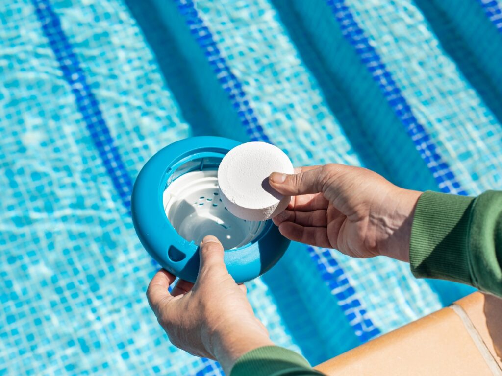 A person places a chlorine tablet into a floating dispenser beside a swimming pool with blue water and tiled edges, helping to avoid one of the most common pool safety mistakes: improper chemical treatment.