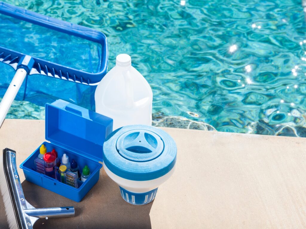 Pool cleaning supplies, including a net, a gallon jug, a chemical test kit, and a floating chlorine dispenser, are arranged next to a swimming pool to help prevent some of the most common pool safety mistakes.
