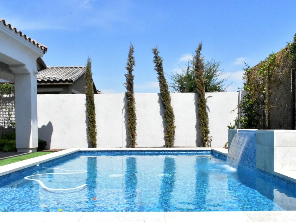 Rectangular backyard swimming pool with a water feature, perfect for New Pool Planning, surrounded by a white wall and tall thin trees, under a clear blue sky.