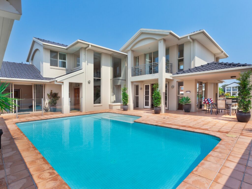Two-story modern house with white walls, large windows, balconies, and a swimming pool featuring the latest pool trends, all surrounded by a tiled patio under a clear blue sky.