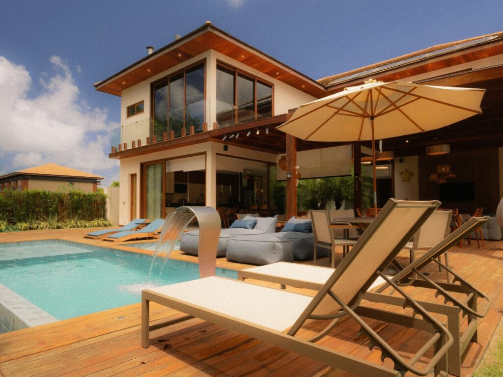 Modern two-story house with wooden deck, lounge chairs, umbrella, and a swimming pool featuring the latest Pool Trends and water feature under a partly cloudy sky.