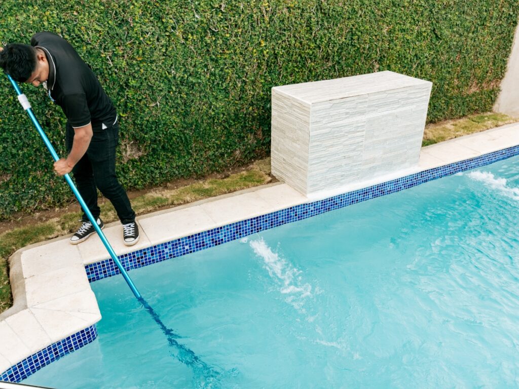 A person in a black shirt and sneakers cleans a swimming pool with a long pole net beside a hedge and a tiled water feature, showcasing the latest Pool Trends in maintenance and design.