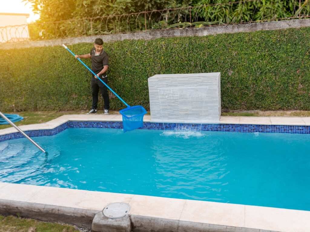 A person performs winter pool maintenance by using a long pool net to clean debris from a backyard swimming pool, standing on the pool edge near a hedge.