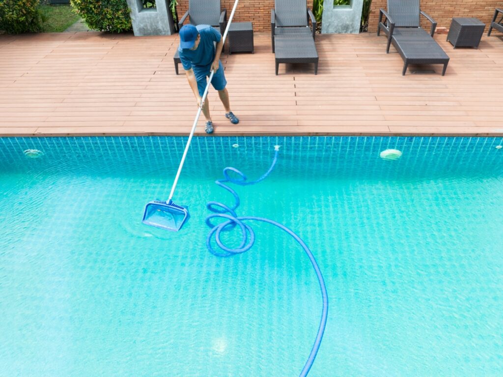 A person in blue clothing uses a pool skimmer to clean a swimming pool beside a deck with several lounge chairs, demonstrating essential winter pool maintenance.