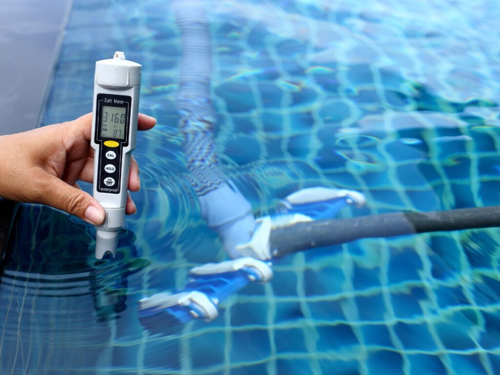 A hand holds a digital water quality tester in a swimming pool, displaying measurements—an essential step in winter pool maintenance. A pool cleaning device is visible underwater on the tiled pool floor.