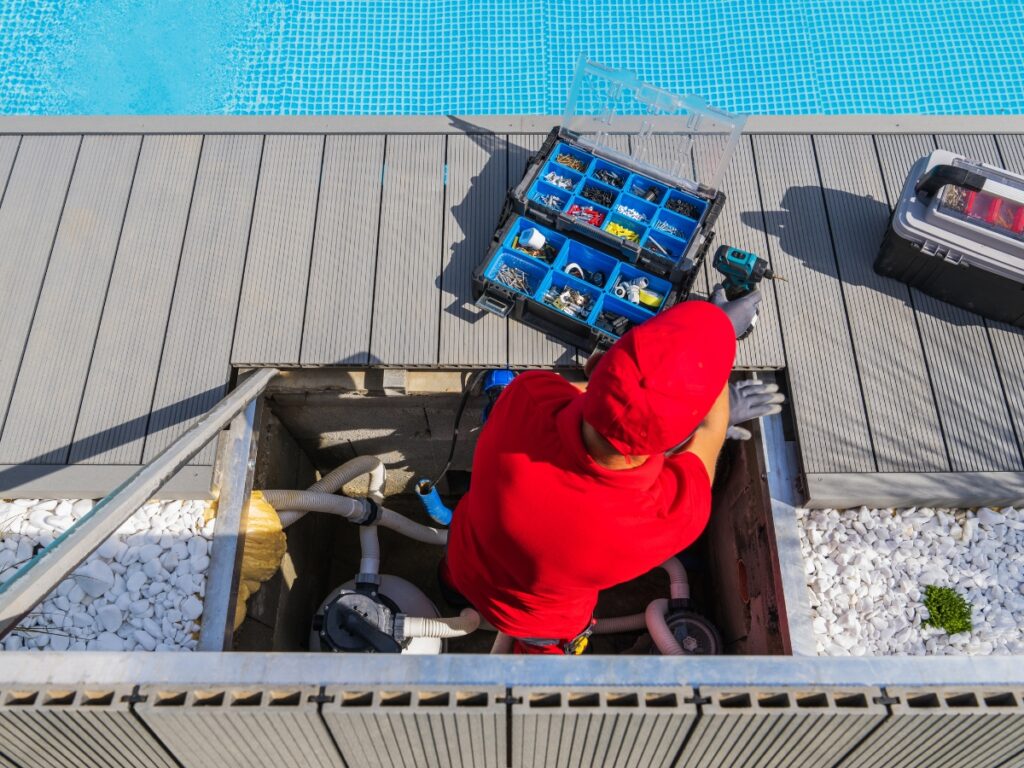 A worker in a red uniform and cap repairs pool equipment in a maintenance hatch beside a swimming pool, with toolboxes nearby, ensuring proper winter pool maintenance.