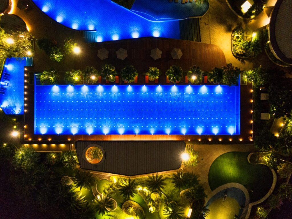 Aerial view of a rectangular illuminated swimming pool at night, featuring stunning design pool lighting, surrounded by lounge chairs, umbrellas, and landscaped greenery.