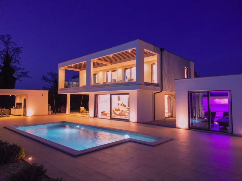Modern two-story house with large windows and a design pool, illuminated by warm lighting at dusk, creating an inviting outdoor ambiance.
