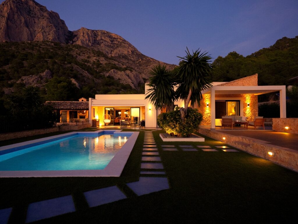 Modern house with large windows and elegant design pool lighting, situated beside a swimming pool with mountains in the background at dusk.