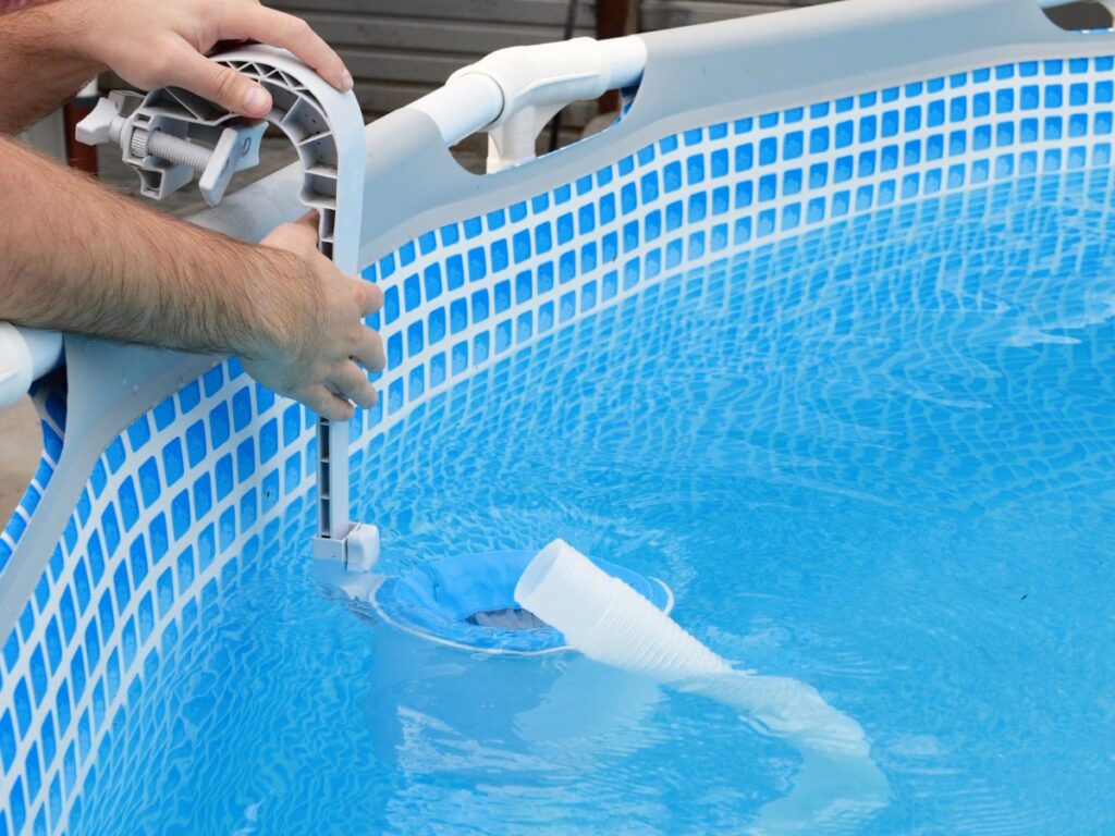 A person installs a pool filter hose connection to the side of a blue above-ground swimming pool, ensuring compliance with local permits regulations.