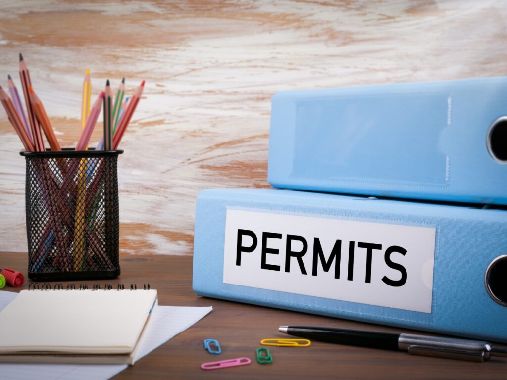 Two blue binders labeled "PERMITS" sit on a desk, ready for reviewing permits regulations—alongside a pencil holder with colored pencils, a notepad, a pen, and paperclips.