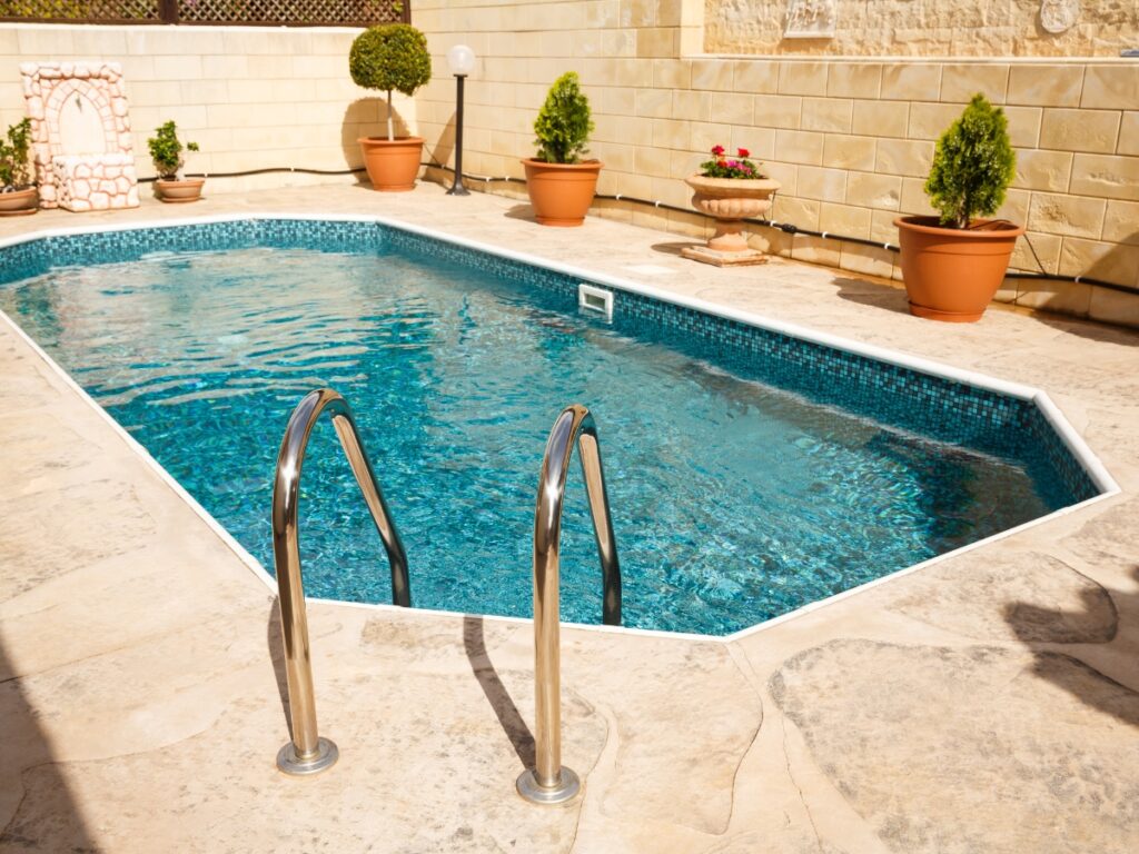 A small outdoor saltwater pool with metal handrails offers gentle swimming and saltwater pool benefits, surrounded by stone tiles and potted plants in a sunny courtyard.