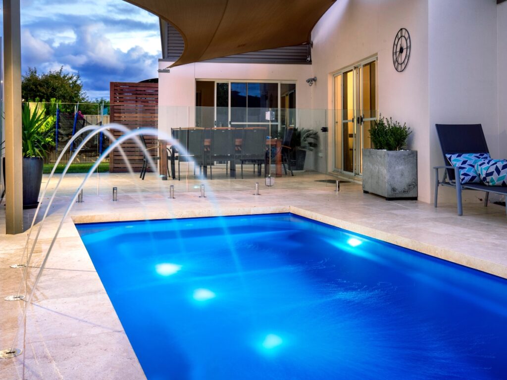 Modern backyard with a small rectangular saltwater pool, water features, illuminated pool lights, a patio seating area, and glass safety fencing—enjoy saltwater pool benefits like gentler water and easy maintenance.