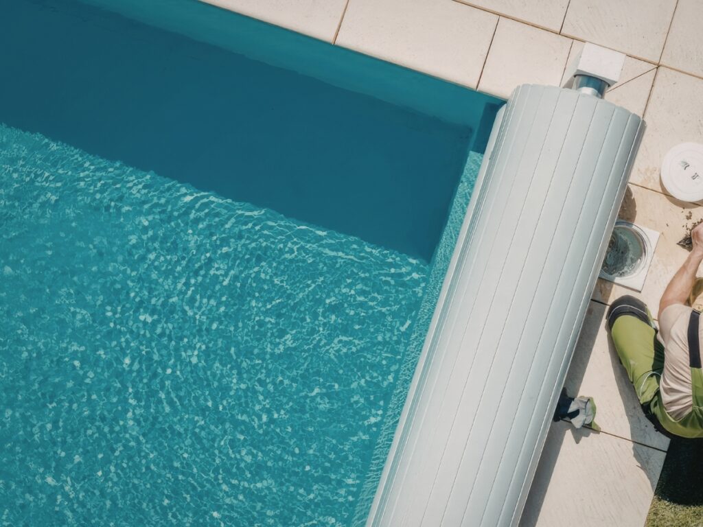 A worker kneels beside a swimming pool, partially covered with one of the latest pool covers, on a tiled pool deck bathed in sunlight.